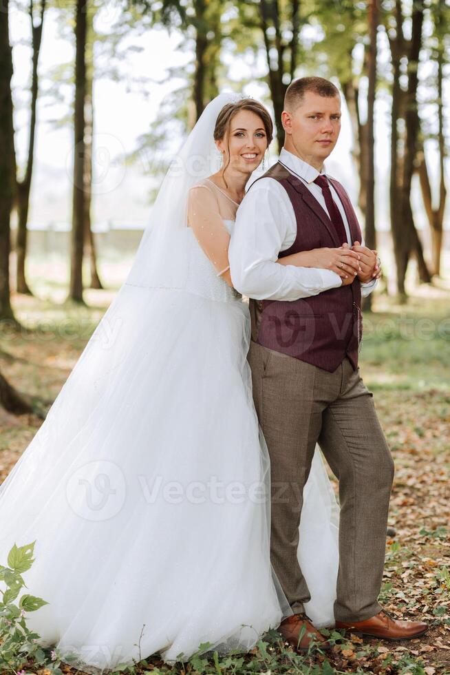 A bride and groom are posing for a picture in a forest. The bride is wearing a white dress and the groom is wearing a red vest. Scene is happy and romantic photo