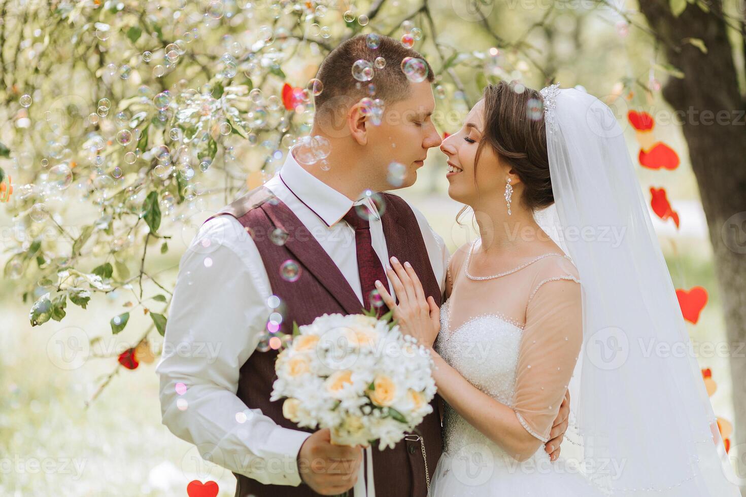 A bride and groom are hugging and kissing in front of a tree. The bride is wearing a white dress and the groom is wearing a red vest photo