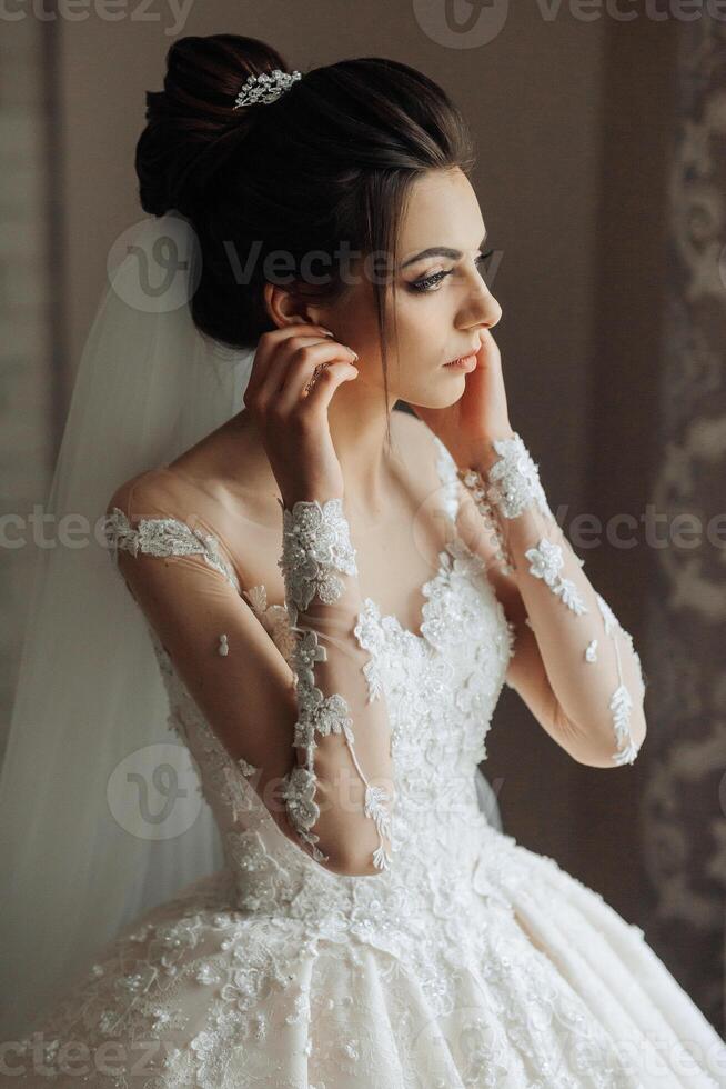 A woman in a wedding dress is looking at her reflection in a mirror. She is wearing a veil and has her hands on her ears. The image has a romantic and elegant mood photo