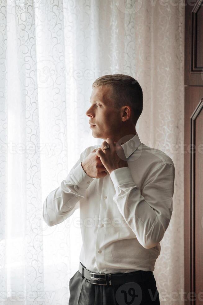 A man in a white shirt is getting ready to tie his tie. He is standing in front of a window, and the light coming in through the window is casting a warm glow on his face photo