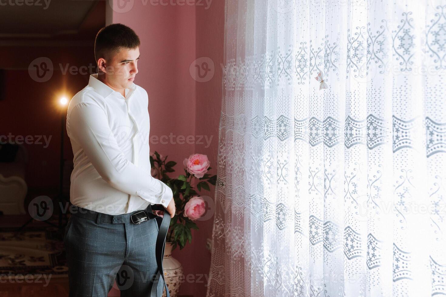 A man in a white shirt stands in front of a window with a floral arrangement in a vase photo