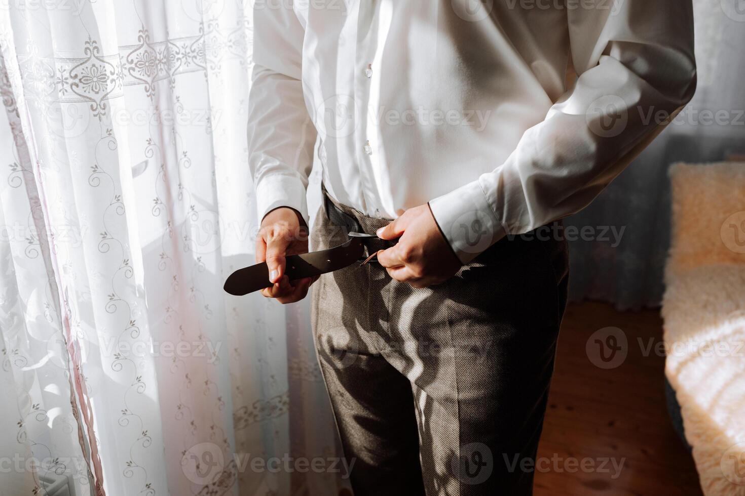 A man in a white shirt is adjusting his belt. Concept of formality and attention to detail, as the man is getting ready for a formal event or occasion. The white shirt and pants suggest a classic photo