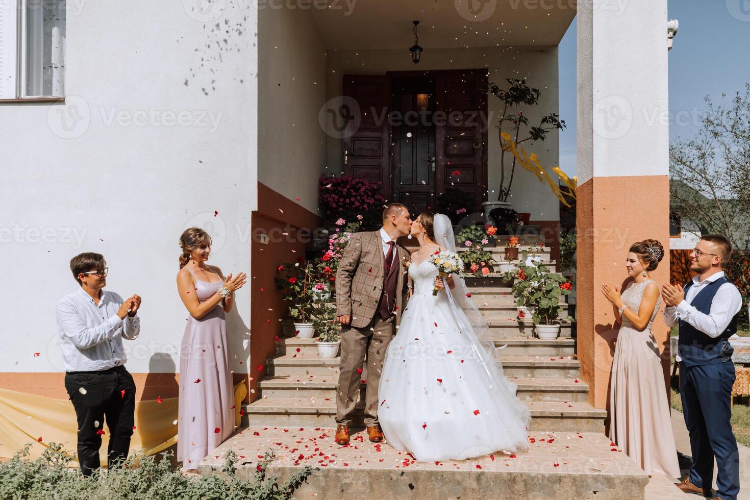 A bride and groom are kissing on a staircase in front of a group of people. The bride is wearing a white dress and the groom is wearing a suit. The people in the background are clapping photo