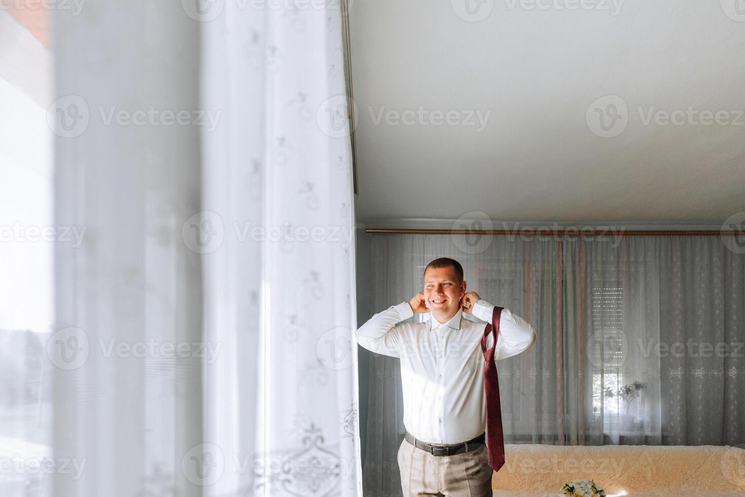 A man in a white shirt and gray pants is standing in front of a window, wearing a red tie. He is listening to music with his earbuds in photo