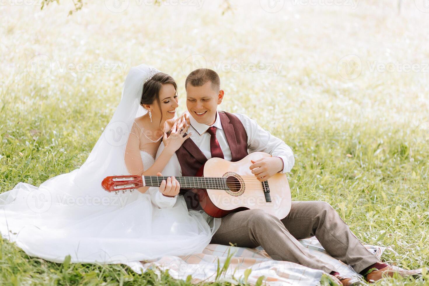 A couple is sitting on a blanket in a grassy field, with a guitar in front of them. The man is wearing a red vest and the woman is wearing a white wedding dress photo