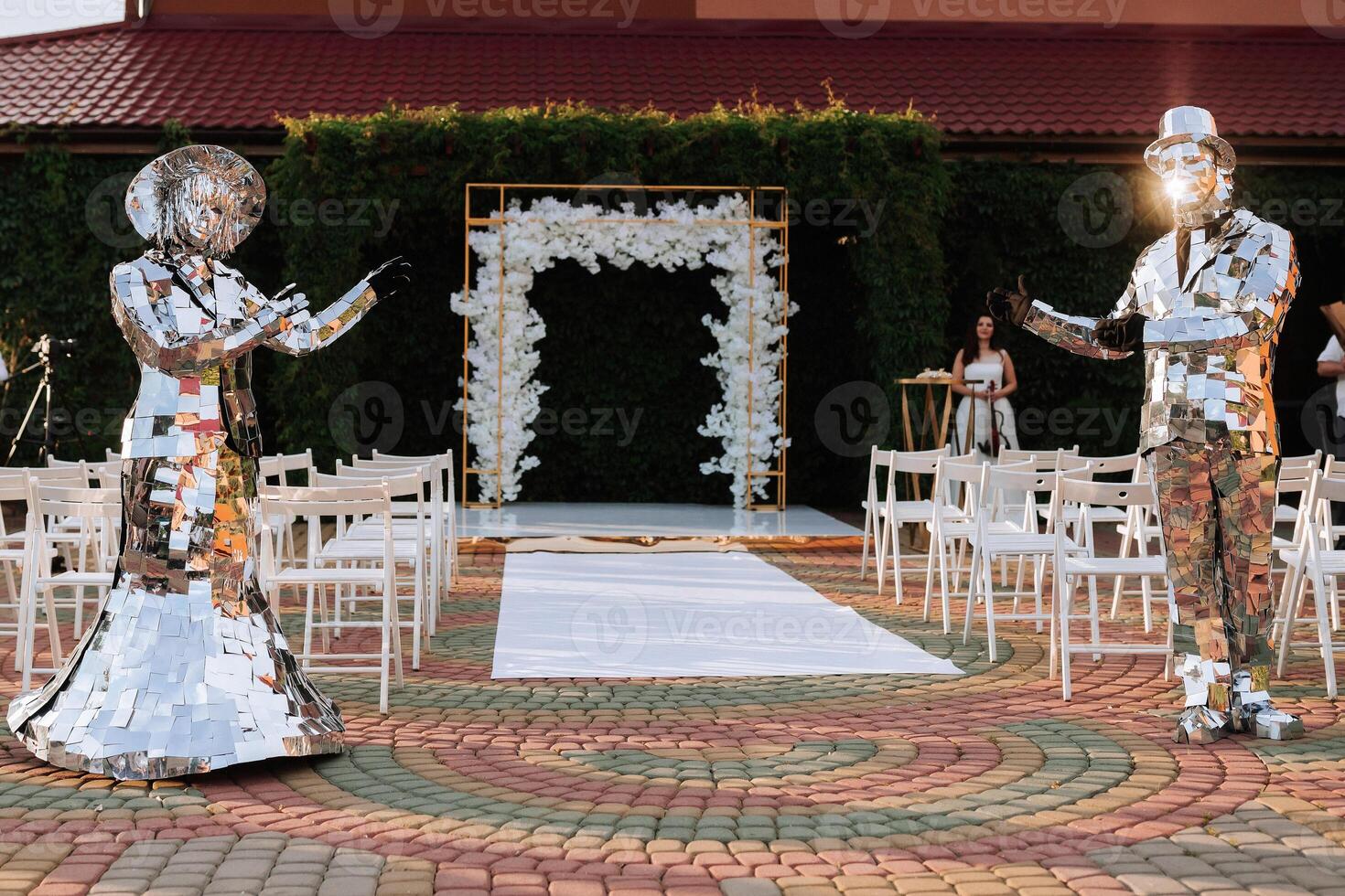 A couple dressed in shiny, metallic costumes stand in front of a white wedding arch. The man is wearing a hat and the woman is wearing a dress. The scene is set in a garden, with chairs photo