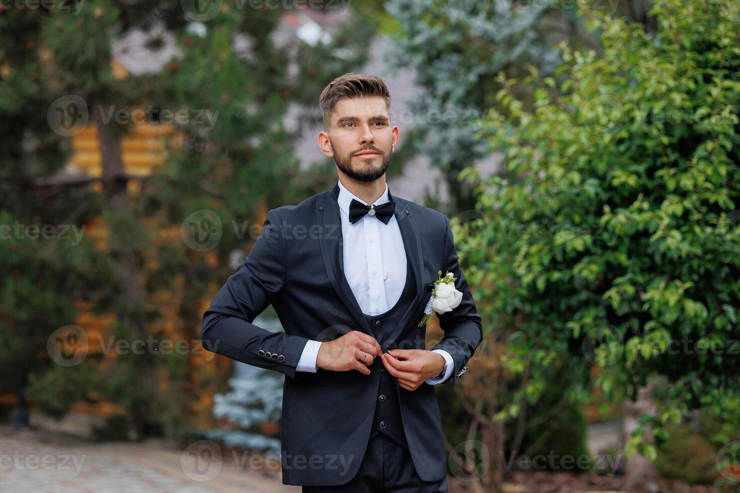 A man in a suit and tie stands in front of a house with a green bush behind him photo