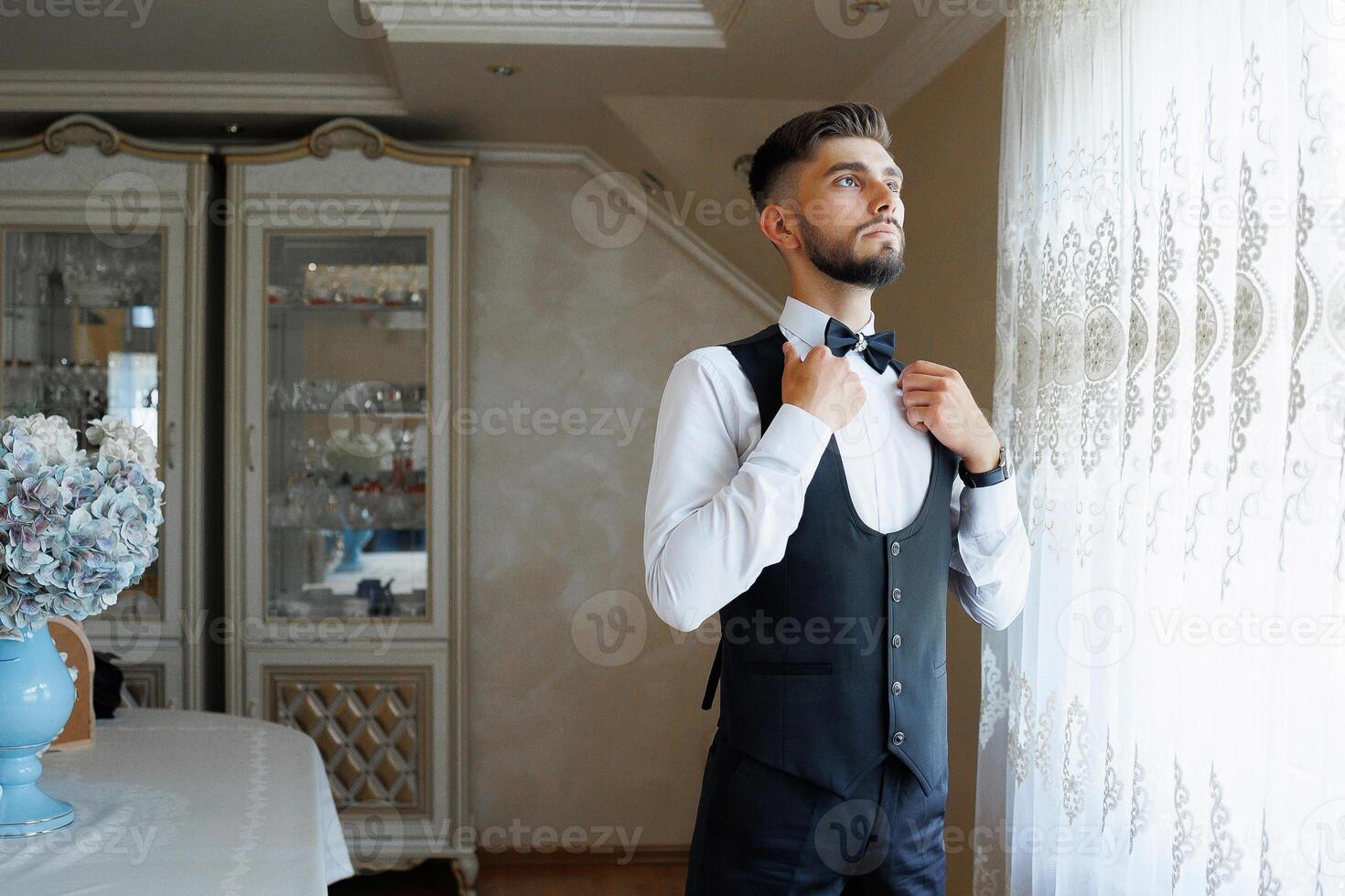 A man in a suit and tie is standing in front of a window, adjusting his tie. Concept of formality and professionalism, as the man is dressed in a suit and tie photo