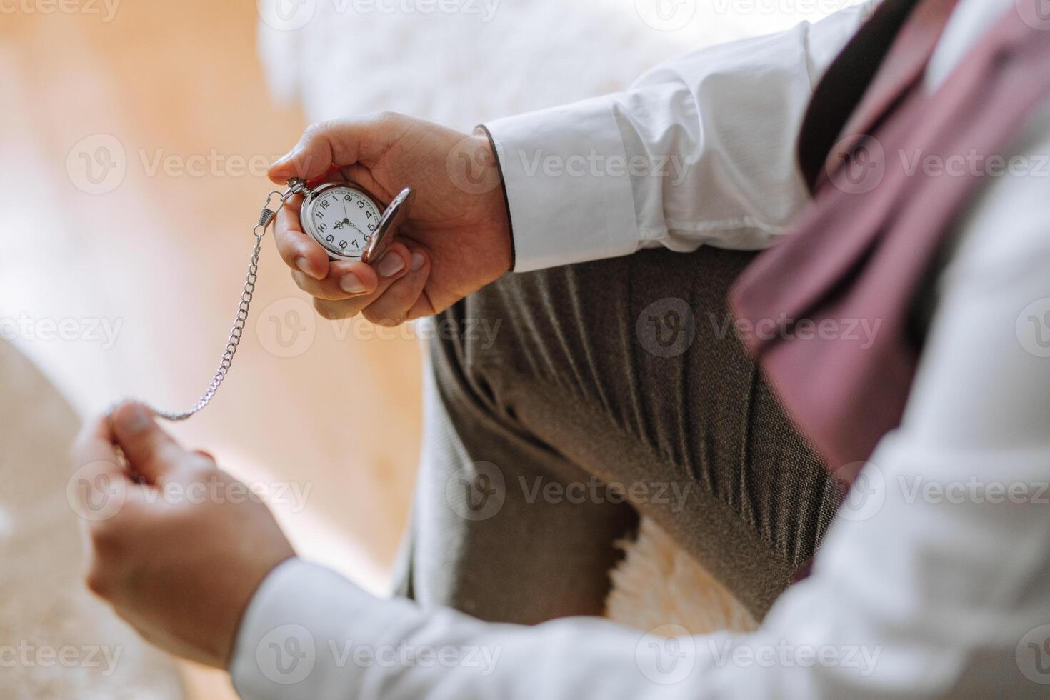 A man is sitting on a rug with a pocket watch in his hand. He is wearing a white shirt and a purple vest photo