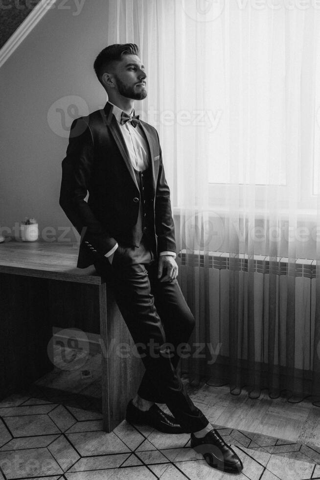 A man in a suit and tie is standing in front of a window. He is wearing a bow tie and has a serious expression on his face photo