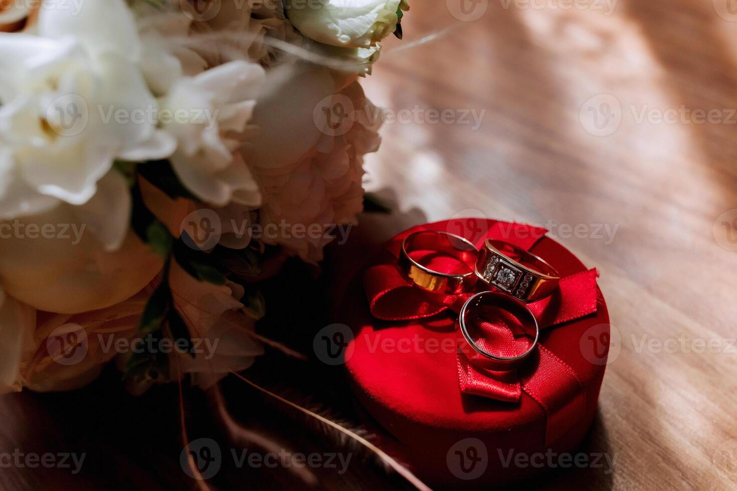 A red box with three rings inside of it. The box is placed on a wooden table. The rings are gold and diamond-studded. The flowers in the background are white photo