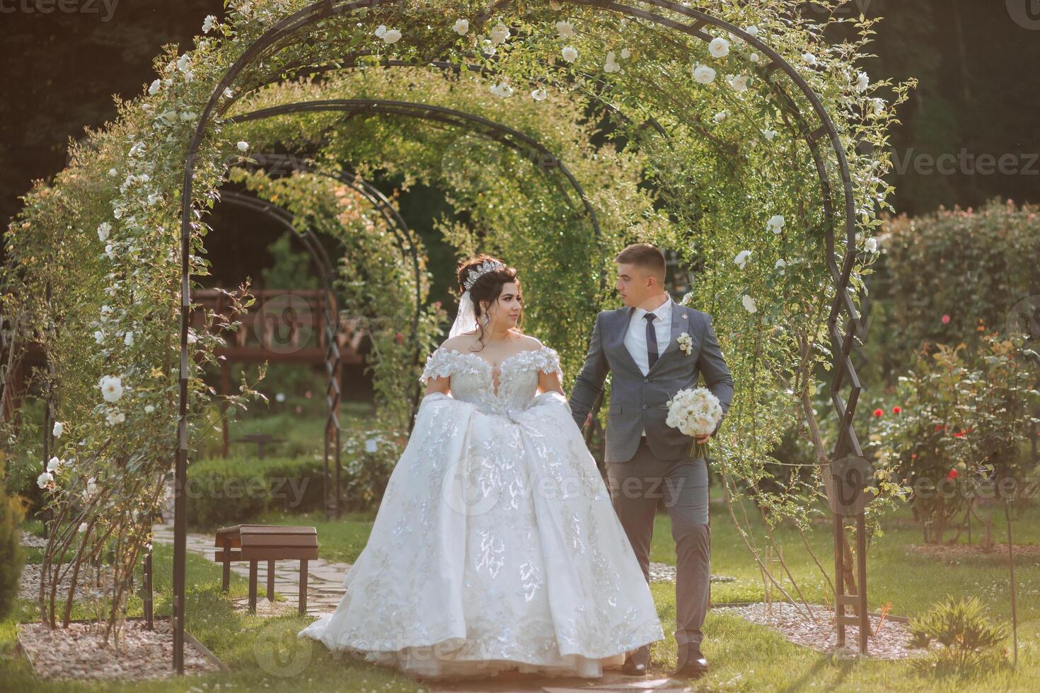 A bride and groom are walking through a garden, with the bride wearing a white dress and the groom wearing a suit photo