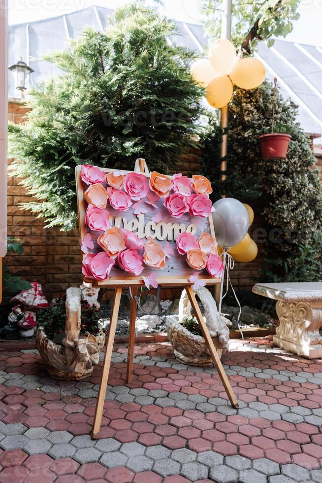 A sign with pink flowers and the word written on it. The sign is on a wooden stand and is surrounded by potted plants photo