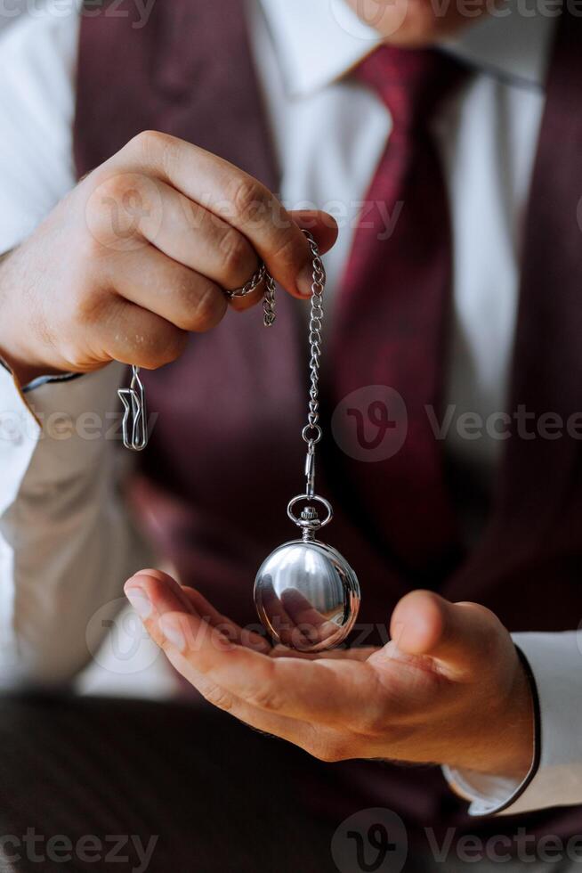 A man is holding a pocket watch in his hand. The watch is silver and has a chain around it. The man is wearing a red vest and a white shirt. Concept of nostalgia and elegance photo
