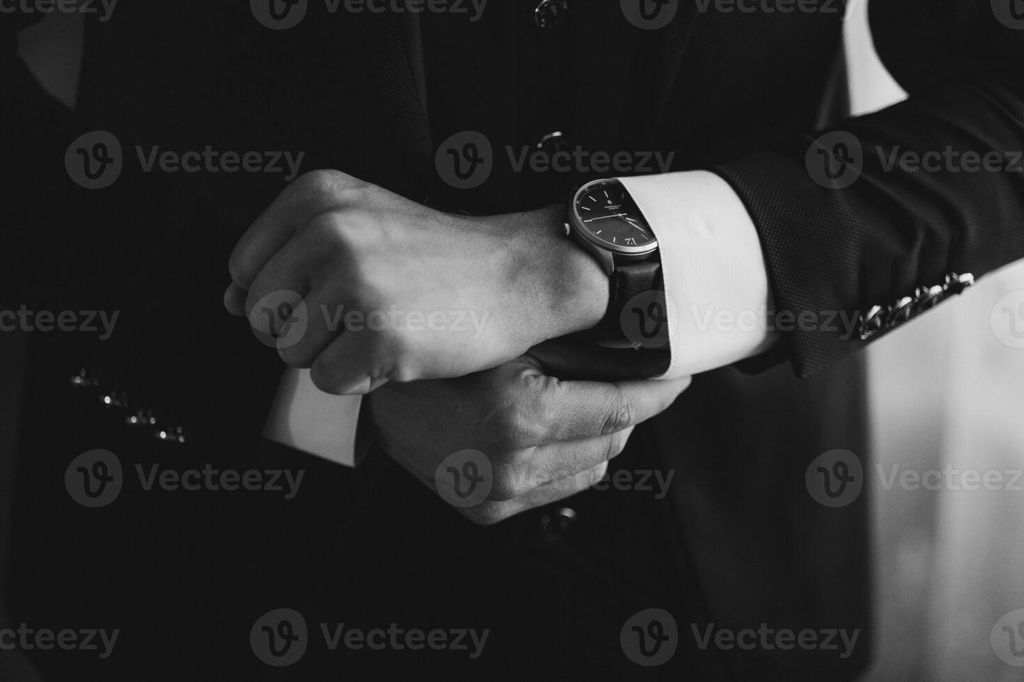 A man is wearing a black suit and is looking at his watch. The watch is black and has a silver band. The man is in a hurry or is checking the time photo