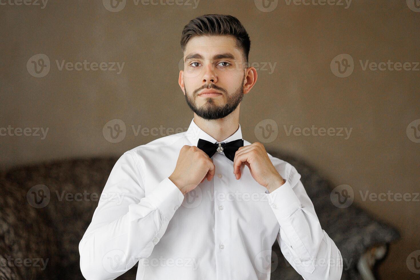 A man in a white shirt and black bow tie is getting ready to go out. He is standing in front of a brown wall photo