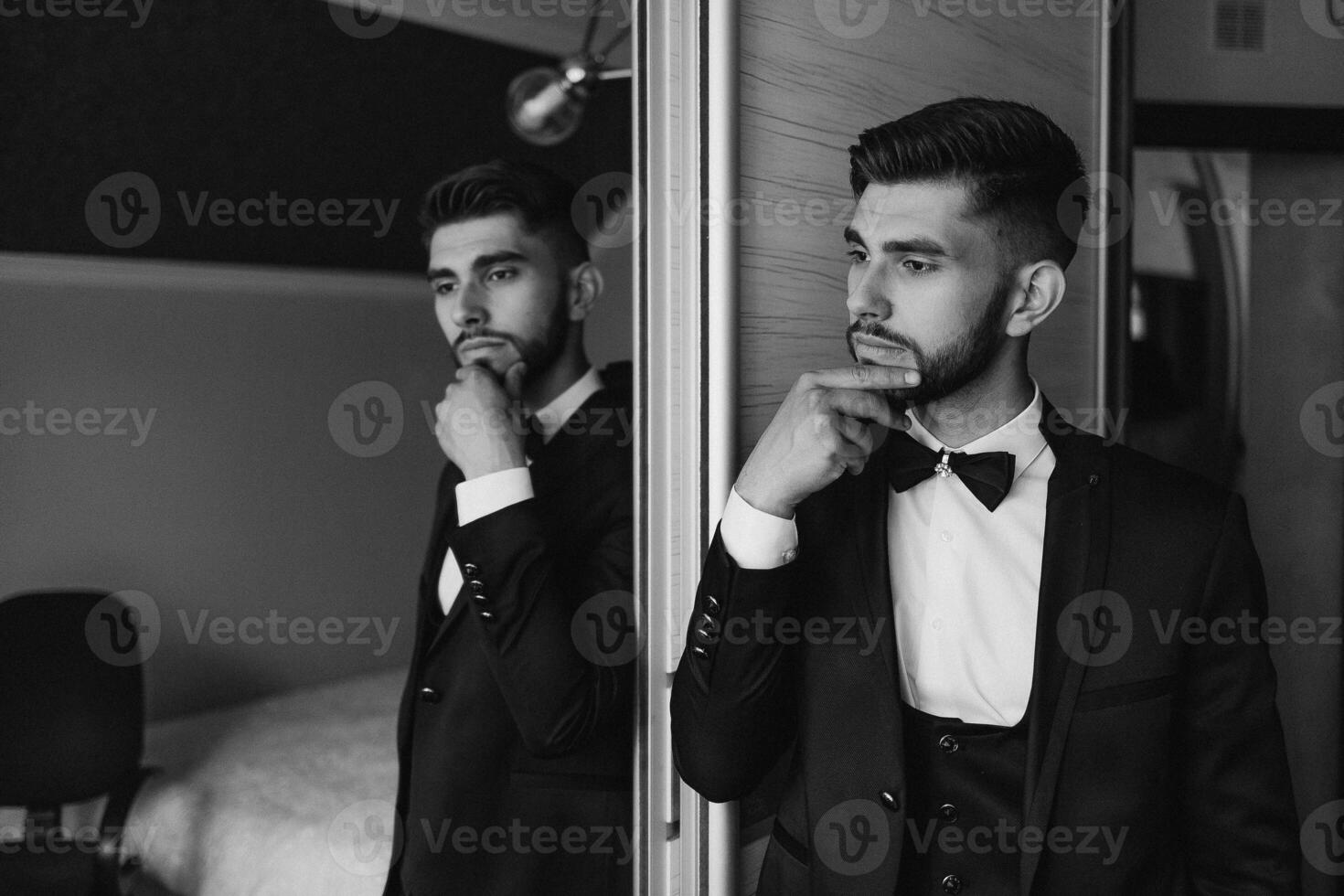 A man in a suit and tie is standing in front of a mirror, looking at his reflection. He is deep in thought, possibly contemplating his outfit or his day ahead. Concept of formality and professionalism photo