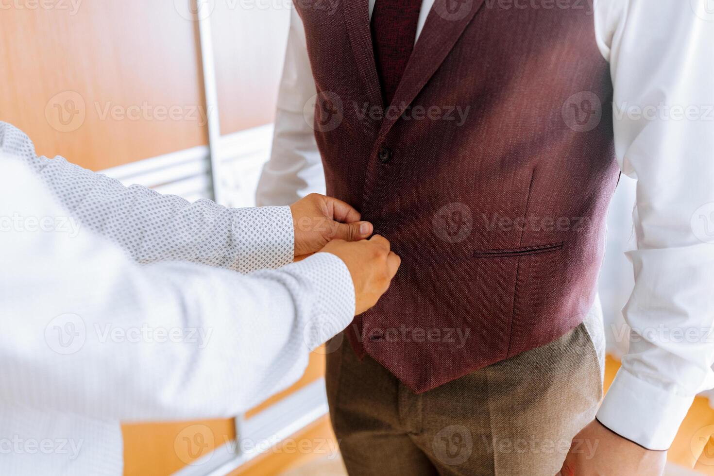 A man is getting dressed with a man helping him. The man is wearing a vest and a tie photo