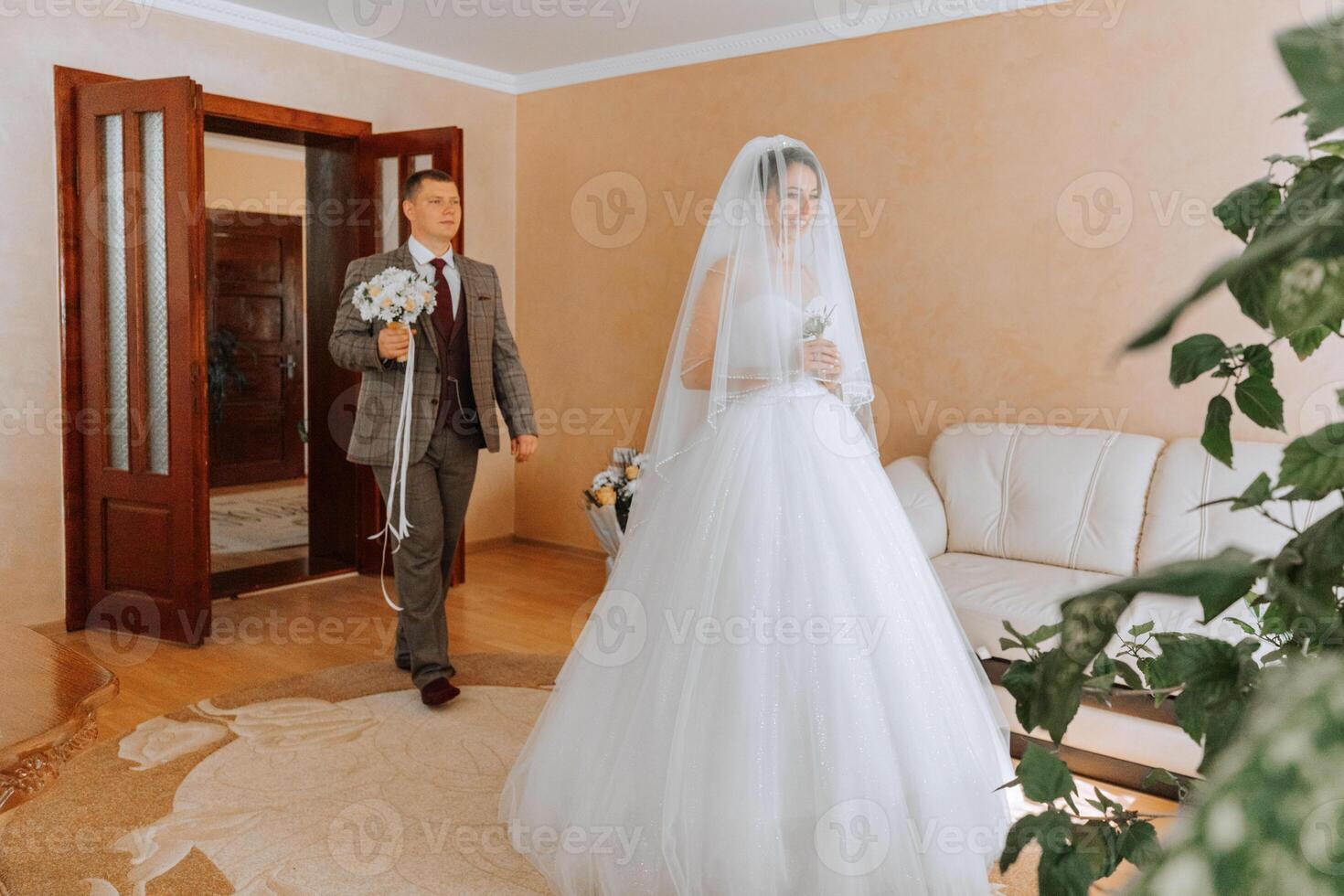 A bride and groom are standing in a living room, with the bride wearing a veil and the groom holding a bouquet photo