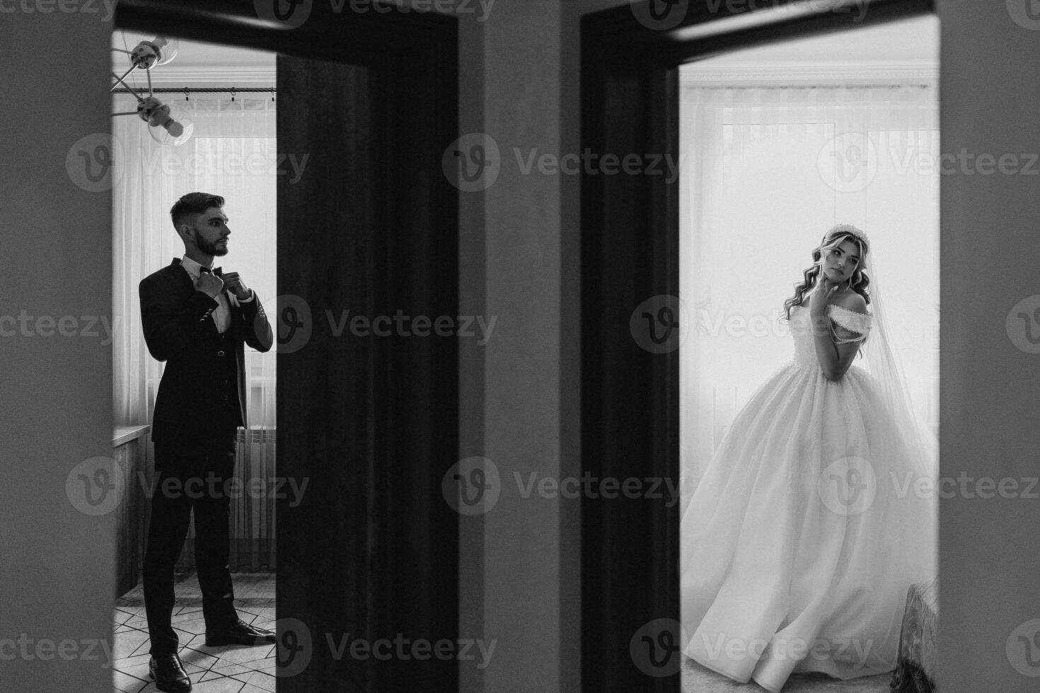 A man and a woman are standing in front of a mirror, with the man wearing a suit and the woman wearing a wedding dress. The man is adjusting his tie, and the woman is looking at him photo