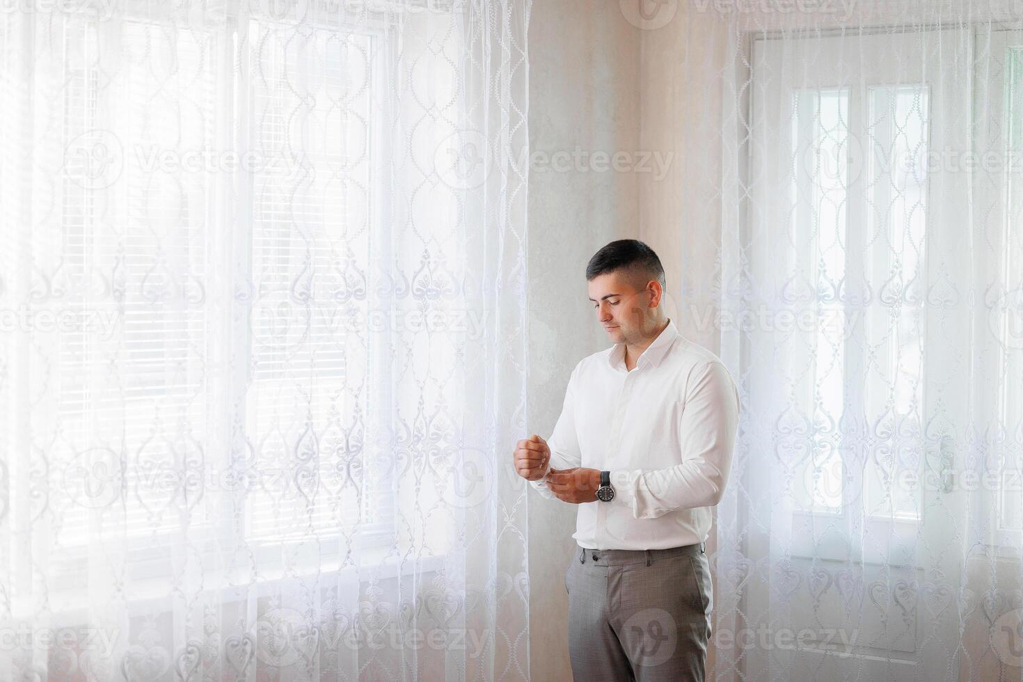 A man in a white shirt is standing in front of a window with a lace curtain. He is adjusting his tie and looking out the window. Concept of formality and attention to detail photo