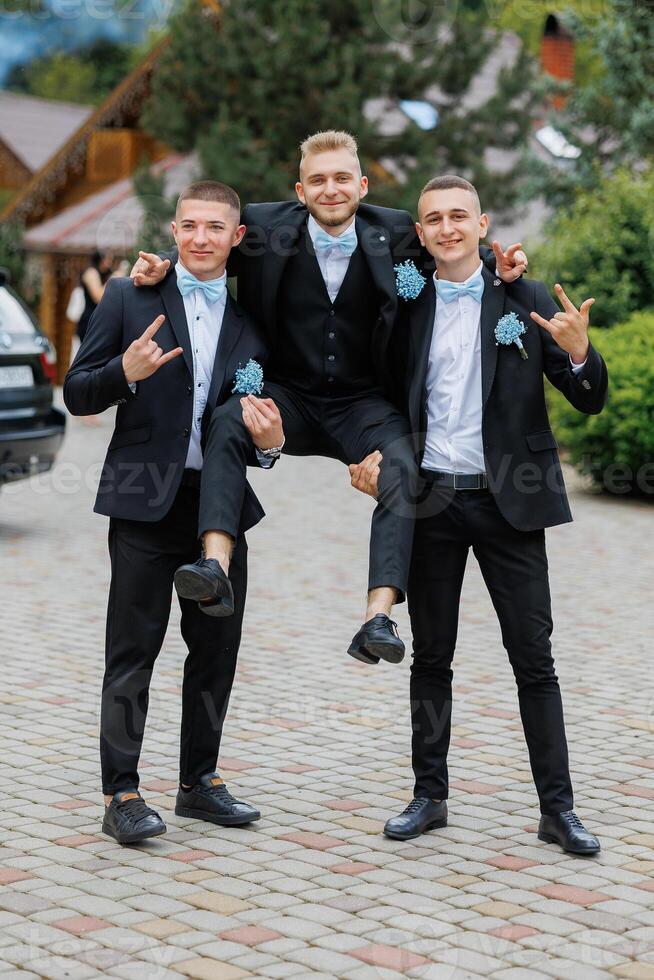 Three men in suits are posing for a picture, with one man holding the other two up. Scene is lighthearted and fun, as the men are dressed up in formal attire and are posing for a photo