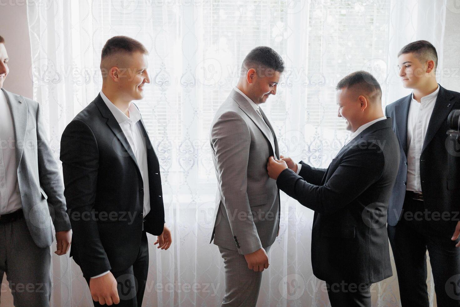 A group of men are getting dressed up for a wedding. One man is helping another man put on his tie photo