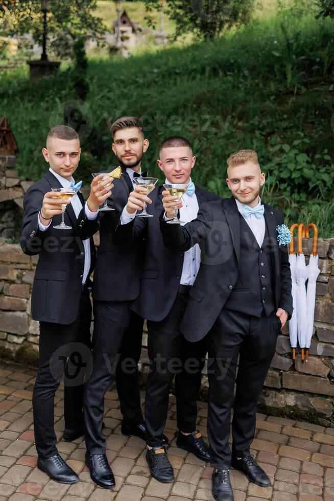 Four men in suits are posing for a photo, each holding a glass of wine. Scene is celebratory and festive, as the men are dressed in formal attire and are toasting to a special occasion photo