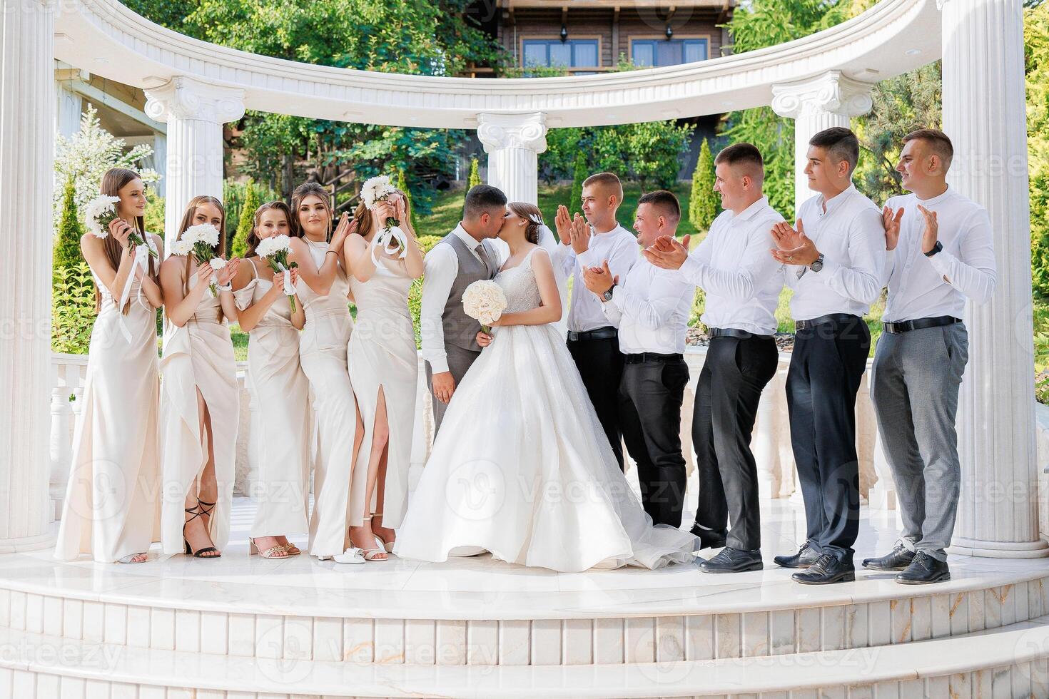 A group of people are posing for a picture, with a bride and groom in the center. The bride is wearing a white dress and the groom is wearing a suit. The group is standing in front of a white archway photo