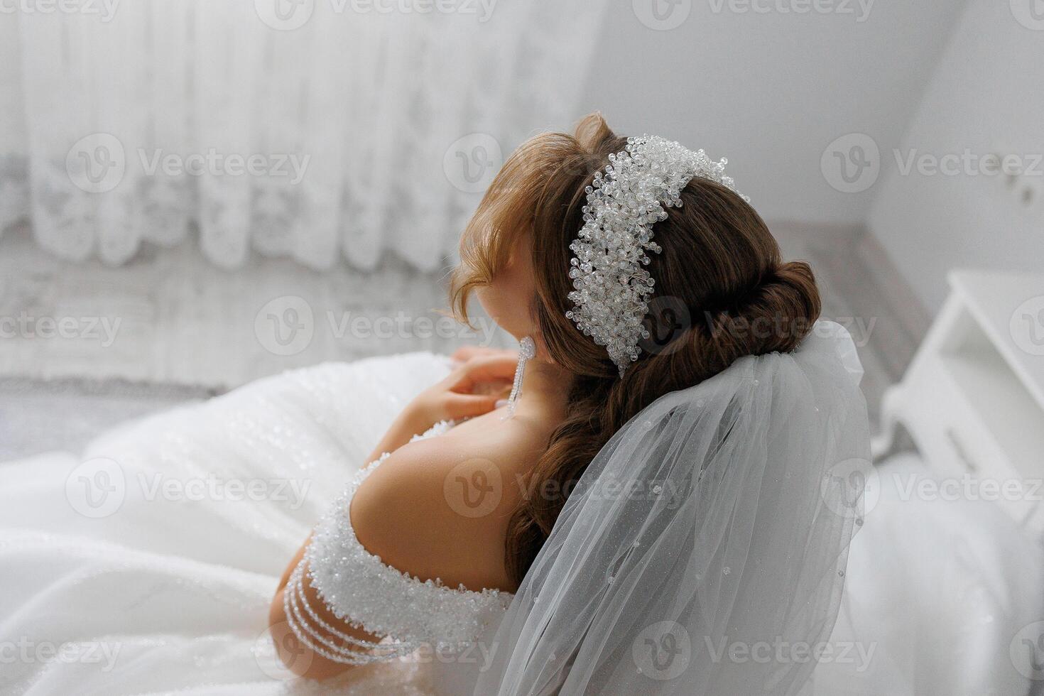 A woman is wearing a wedding dress and a veil. She is sitting on a bed. The image has a blurry effect, which gives it a dreamy and romantic mood photo