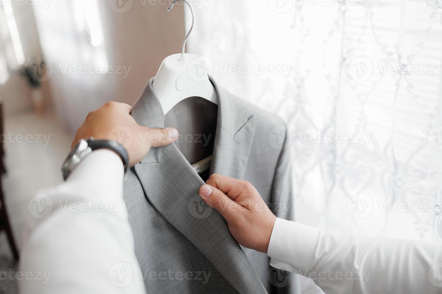 A man is looking at a jacket hanging on a hanger. He is wearing a watch and a white shirt photo