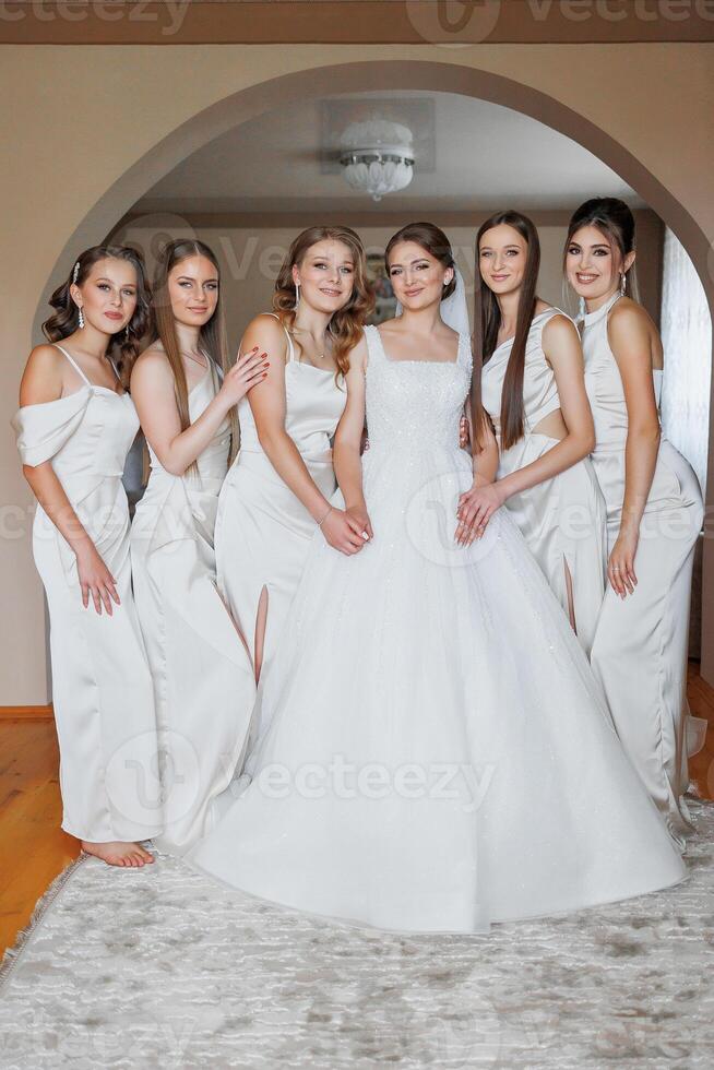 A group of young women are posing for a picture in front of a bride's wedding dress. The bride is wearing a white gown and is surrounded by her five bridesmaids. Scene is joyful and celebratory photo