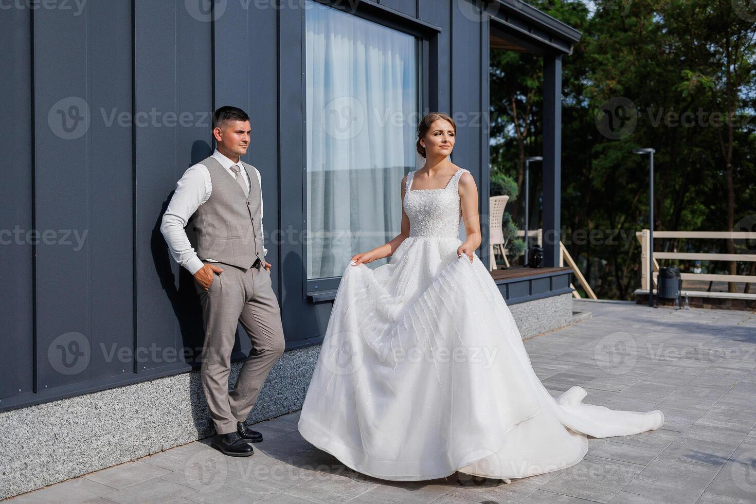 A bride and groom stand outside a house, the bride holding a dress. Scene is happy and celebratory, as the couple is getting ready for their wedding photo