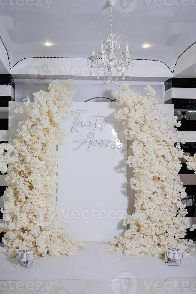 A white archway with the words written on it. The archway is decorated with white flowers and is surrounded by a white backdrop. The scene is elegant and romantic photo