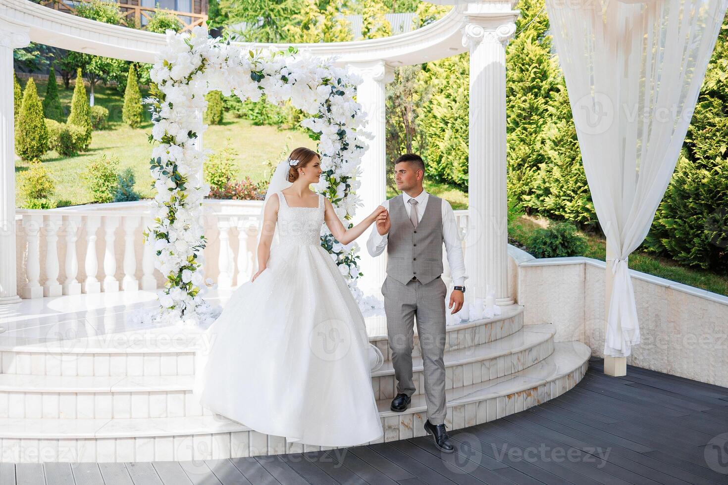 A bride and groom are walking up the steps of a white archway. The bride is wearing a white dress and the groom is wearing a gray suit. They are holding hands photo
