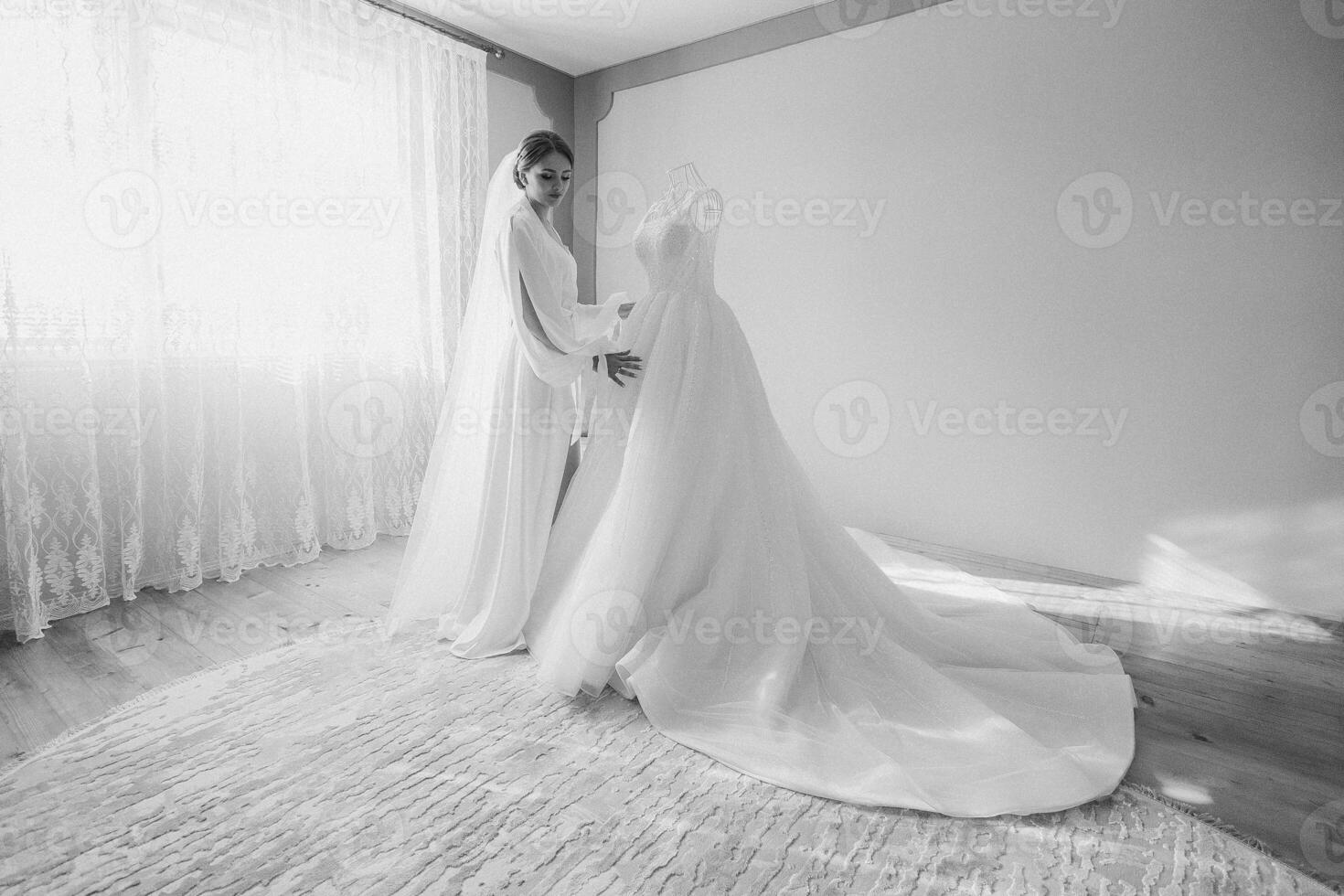 A bride is getting ready for her wedding and is adjusting her dress. The bride is standing in front of a white dress and a white curtain photo