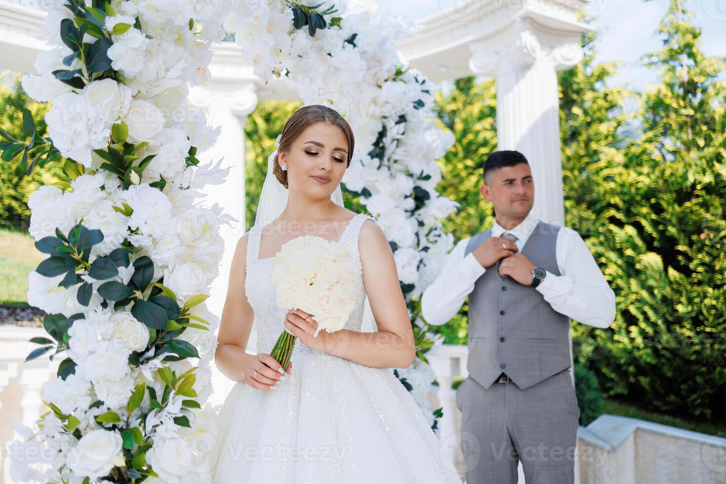 A bride and groom are standing in front of a white archway. The bride is holding a bouquet of flowers and the groom is adjusting his tie. The scene is likely a wedding ceremony or reception photo