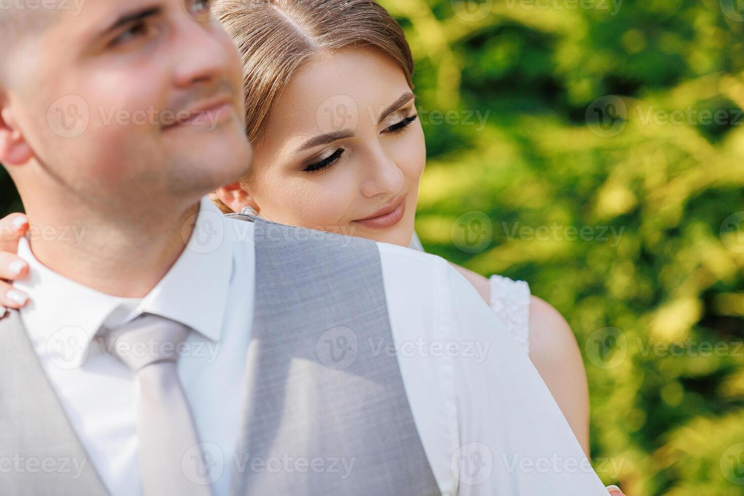 A bride and groom are hugging each other in a lush green field. The bride is wearing a white dress and the groom is wearing a gray suit. Scene is one of love and happiness photo