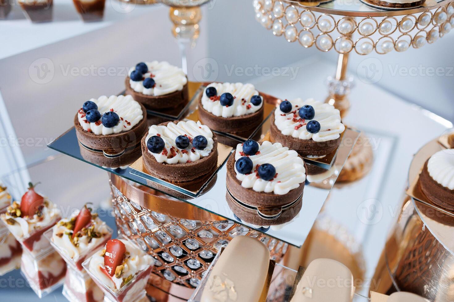 A table with a variety of desserts, including blueberry tarts and strawberry shortcakes. The desserts are arranged on a glass display case, which is surrounded by a gold frame photo