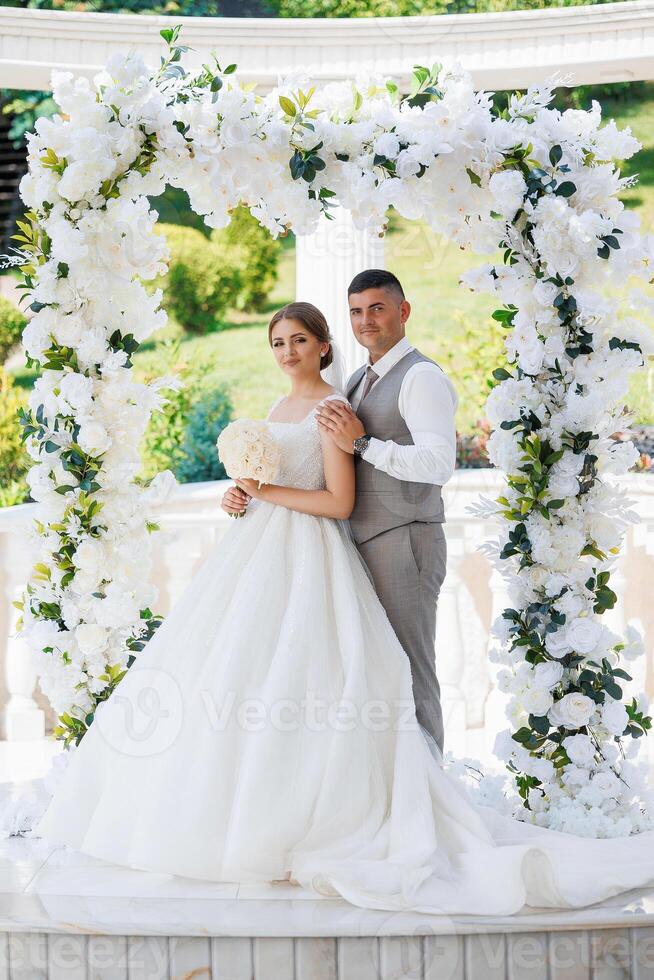 A bride and groom are posing for a picture under a white arch. The bride is holding a bouquet and the groom is wearing a vest. The scene is set in a garden or a park, with a white arch as the backdrop photo