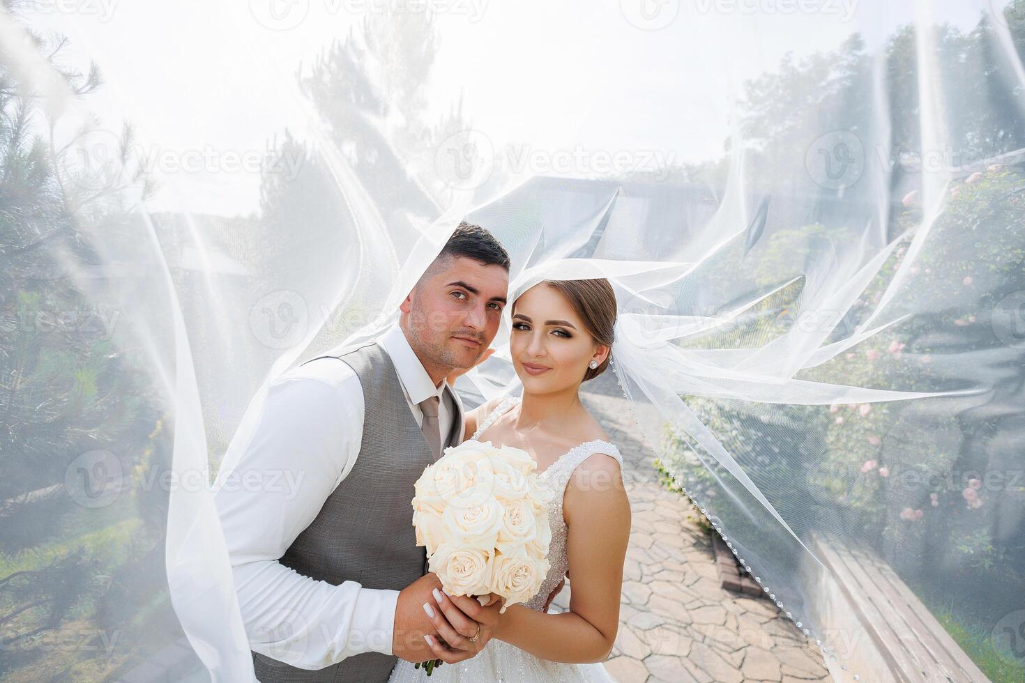A bride and groom are posing for a picture with a white veil. The bride is holding a bouquet of flowers photo