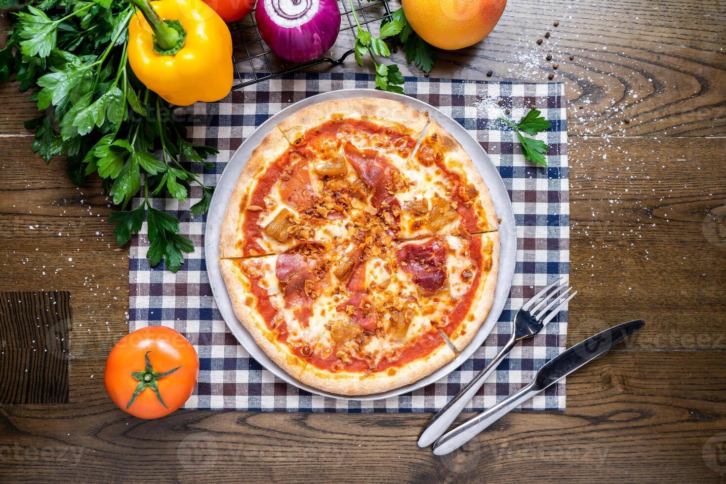 Hawaiian Pizza with tomato, onion, capsicum, coriander, knife and fork served in dish isolated on napkin top view of italian fastfood on wooden table photo