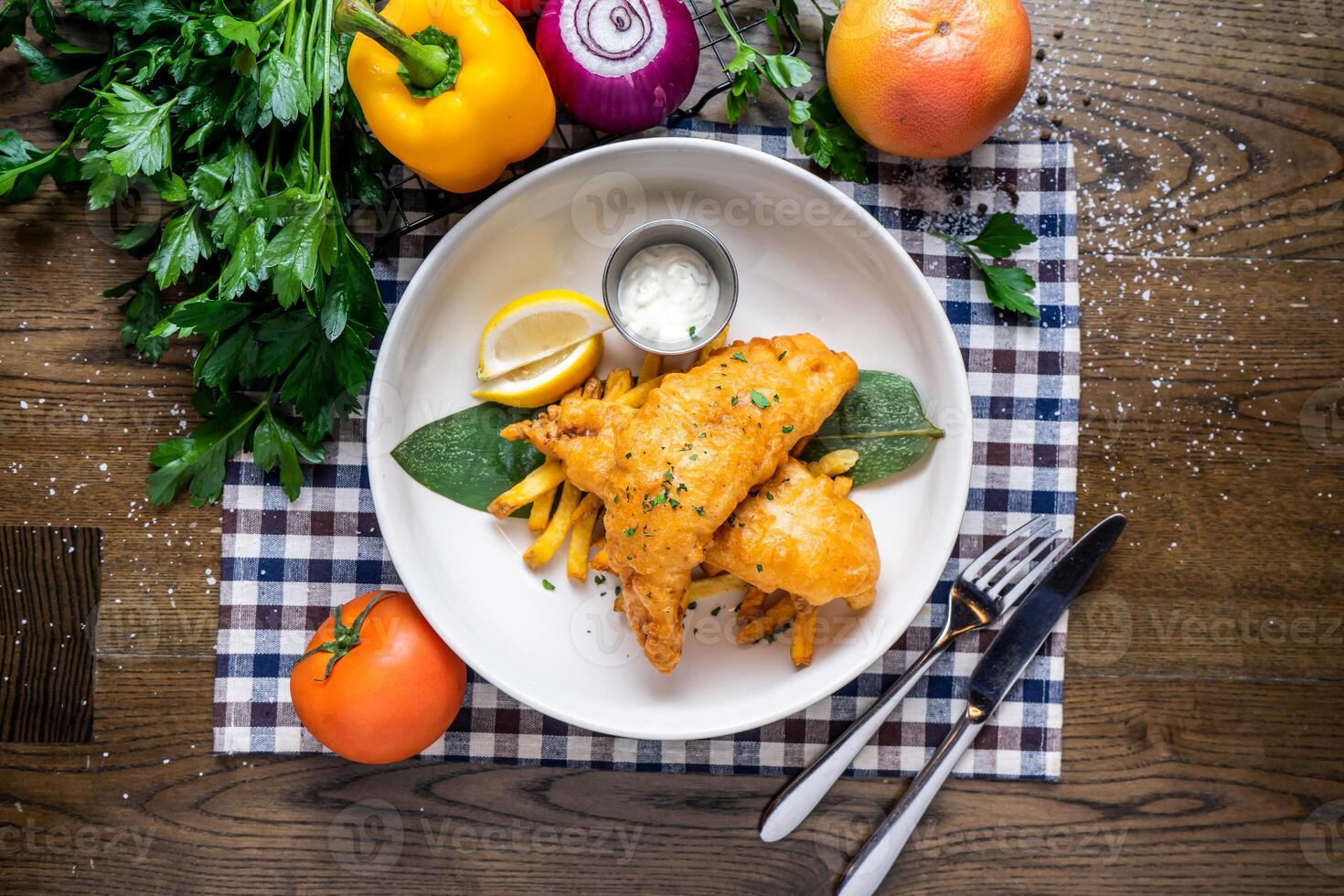 Fried Fish And Chips with lemon, dip, tomato, onion, capsicum, coriander, knife and fork served in dish isolated on napkin top view of italian fastfood on wooden table photo