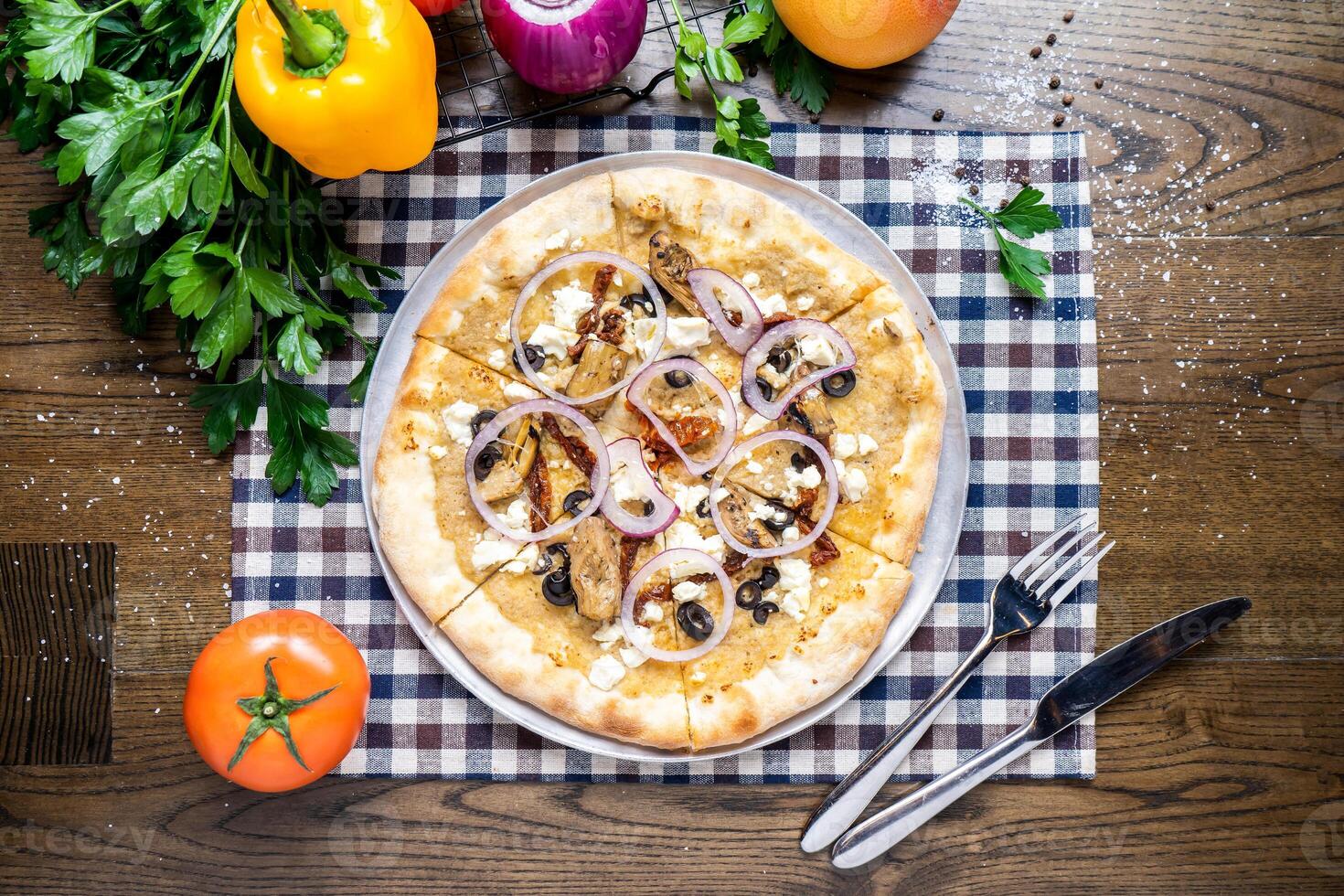 Mediterranean Pizza with black olives, meat, tomato, onion, capsicum, coriander, knife and fork served in dish isolated on napkin top view of italian fastfood on wooden table photo