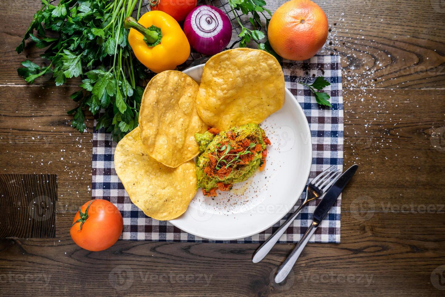 Smash Avocado Bowl with nachos, tomato, onion, capsicum, coriander, knife and fork served in dish isolated on napkin top view of italian fastfood on wooden table photo
