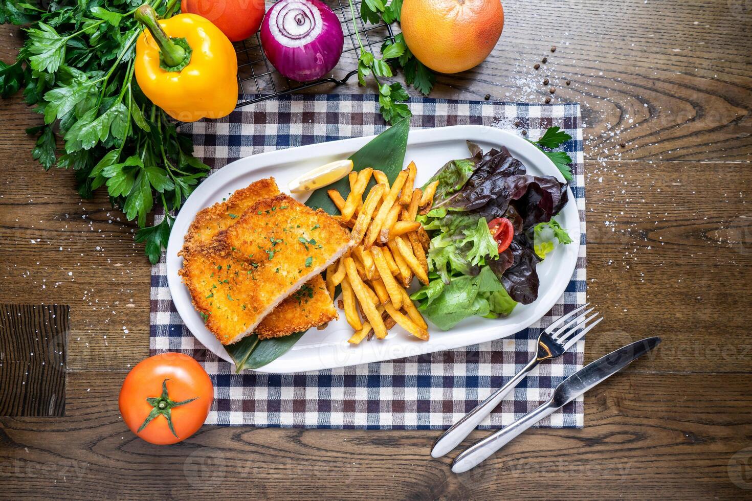Chicken Schnitzel with french fries, salad, tomato, onion, capsicum, coriander, knife and fork served in dish isolated on napkin top view of italian fastfood on wooden table photo