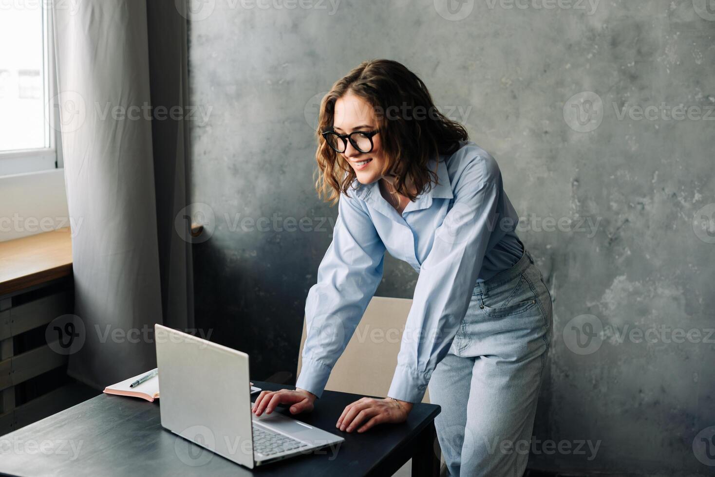 Professional woman with glasses sitting at a desk, working on a laptop in a bright office environment. Perfect for themes related to remote work, online teaching, or modern business settings photo