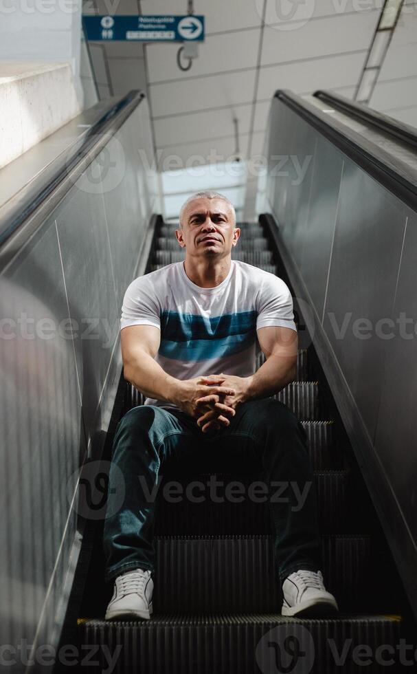 A man in a white shirt and blue jeans sits on a set of escalators photo