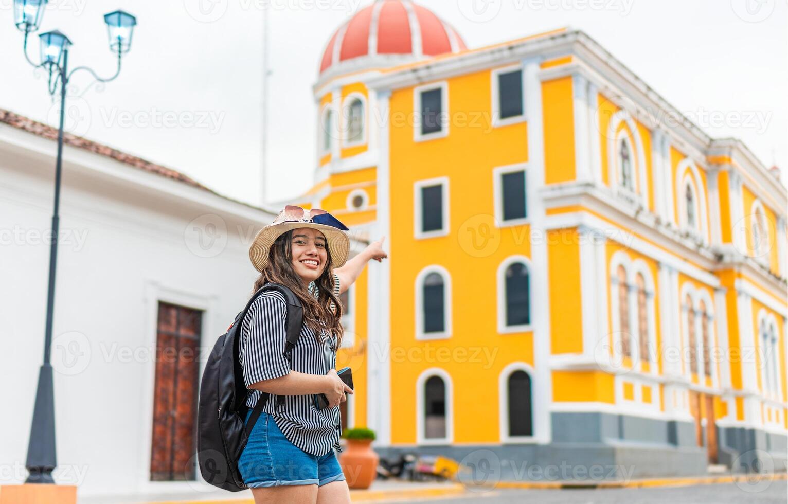 Smiling traveler woman with backpack pointing to a cathedral in a tourist square. Happy tourist girl exploring the city of Granada, Nicaragua photo