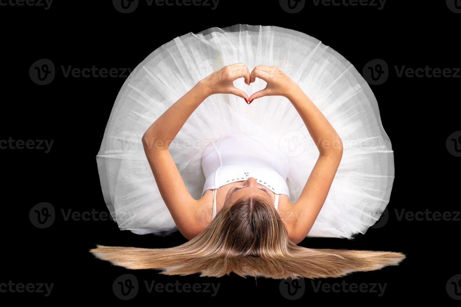 ballerina in a white dress lies on the ground and shows a heart with her hands, portrait on a black background photo